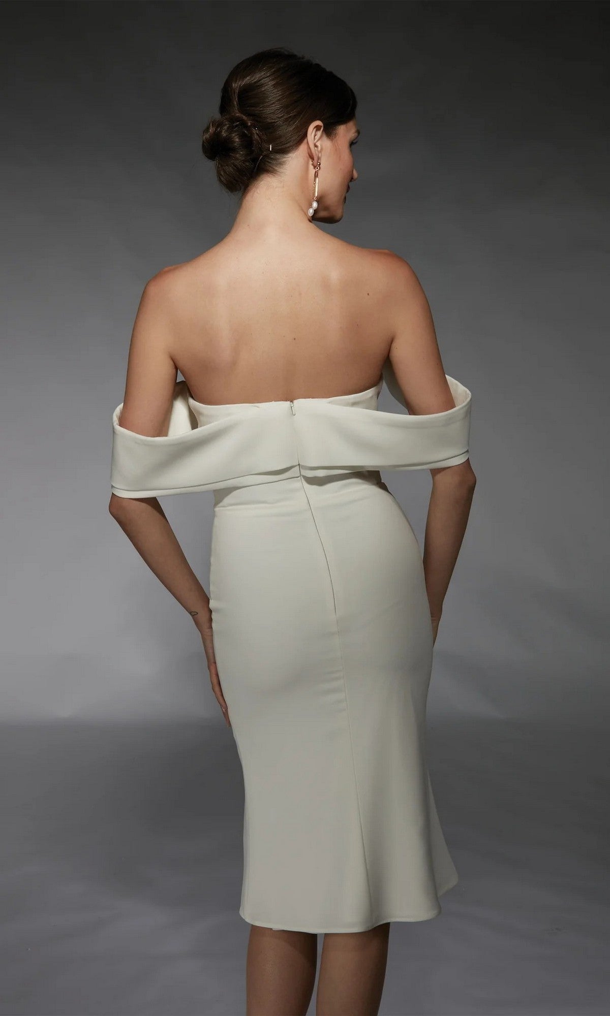 A woman with her hair in a low bun faces away, wearing the Alyce 70055 knee length semi-formal dress, set against a gray background.